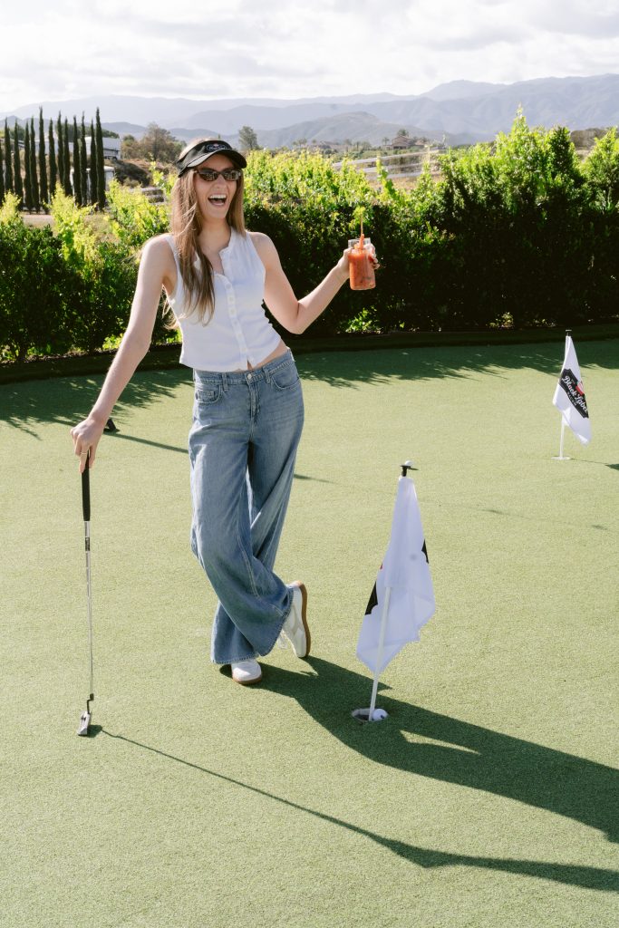 A smiling editor holds a branded drink and a putter on a putting green during a CBC HOUSE Program activity, with branded flags, manicured hedges, and a sweeping mountain and vineyard backdrop behind her.