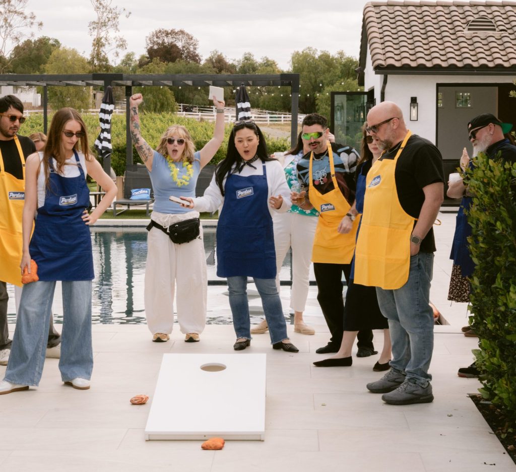Editors and brand representatives wearing Perdue-branded blue and yellow aprons play cornhole poolside at a CBC PR HOUSE Program activation, with a vineyard visible in the background.