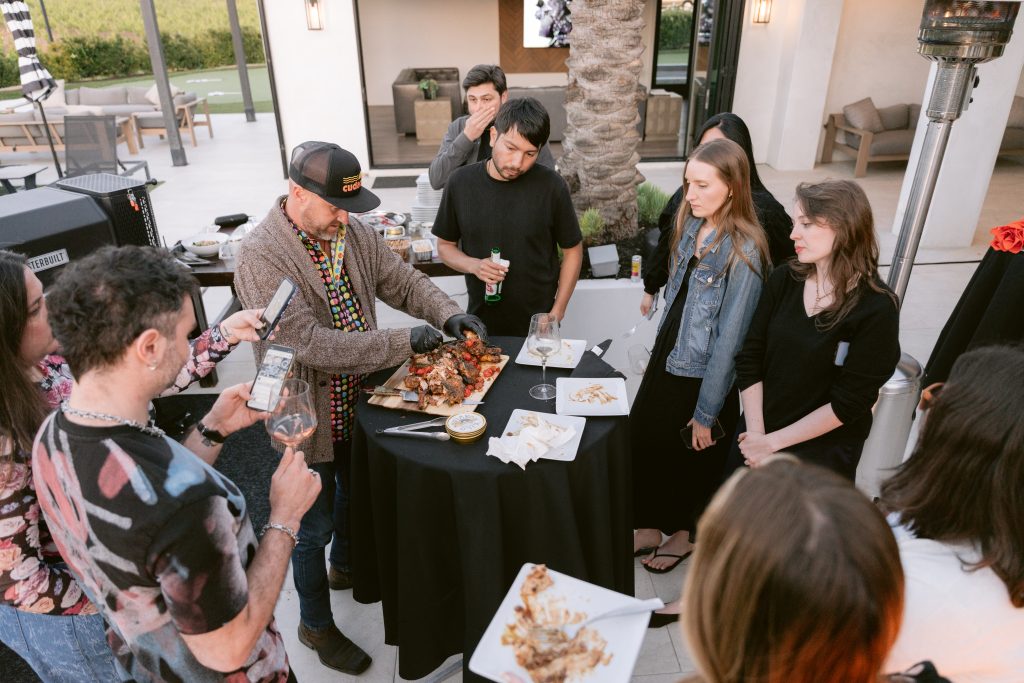A chef in a colorful patterned jacket carves and plates grilled meat at an outdoor evening reception during a CBC HOUSE Program, as a group of editors gathers around the table to watch, drinks in hand, on a patio with palm trees in the background.