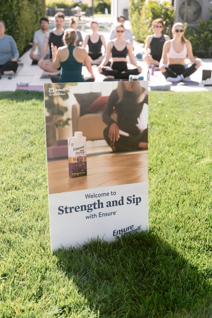 An Ensure "Strength and Sip" branded sign sits in the foreground on a sunlit lawn as editors participate in an outdoor yoga session during a CBC HOUSE Program activation, seated in meditation pose on mats behind it.