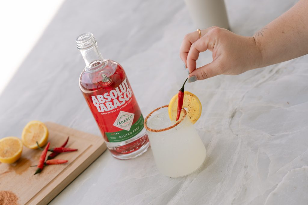 A hand garnishes a spicy cocktail with a red chili pepper, set alongside a bottle of Absolut Tabasco chili pepper-flavored vodka, sliced lemons, and fresh peppers on a white marble surface.