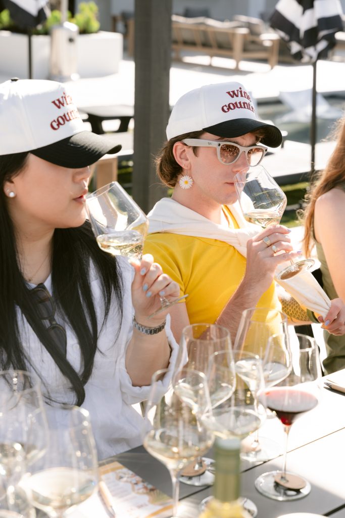 Two editors wearing "Wine Country" branded caps sip white wine at an outdoor tasting table during a CBC HOUSE Program, sunlight catching their glasses in a vineyard setting.