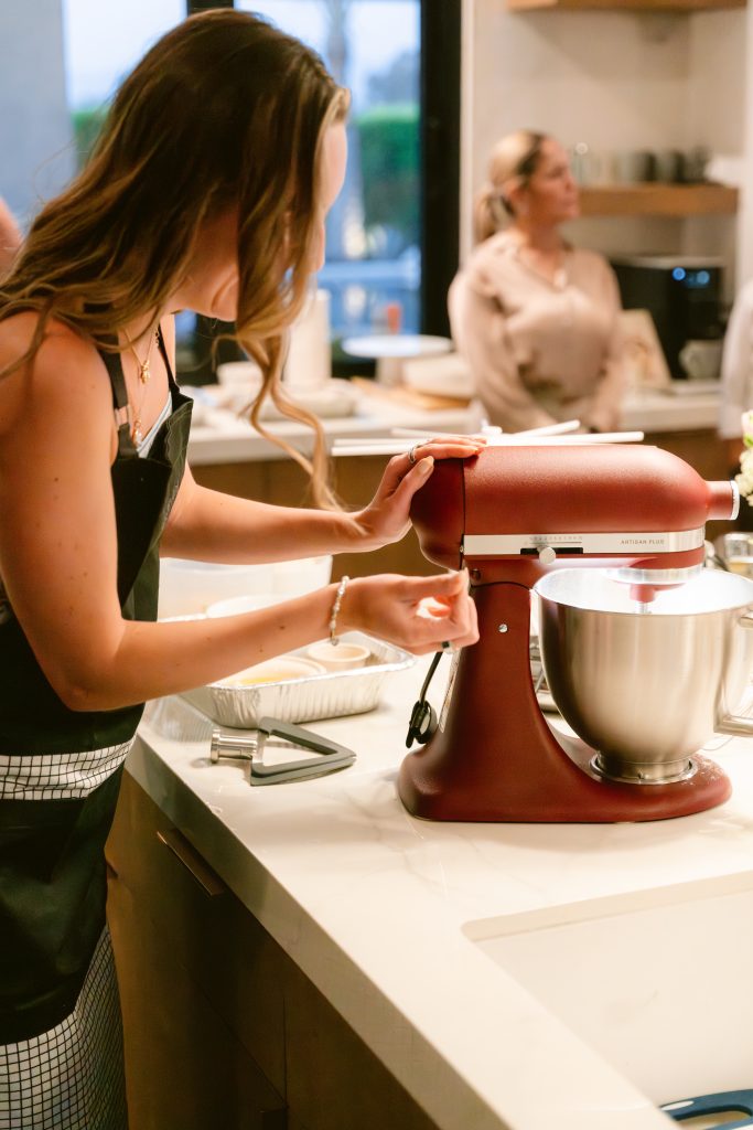 An editor in a dark apron uses a red KitchenAid stand mixer during a hands-on cooking activity at a CBC HOUSE Program, with another participant visible in the background of a well-appointed kitchen.