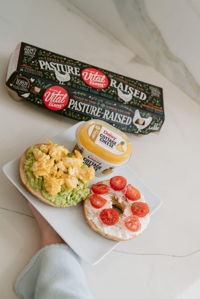 A hand holds a white plate with two open-faced bagels — one topped with scrambled eggs and avocado, one with cottage cheese and cherry tomatoes — alongside a carton of Vital Farms pasture-raised eggs and a container of Daisy cottage cheese.