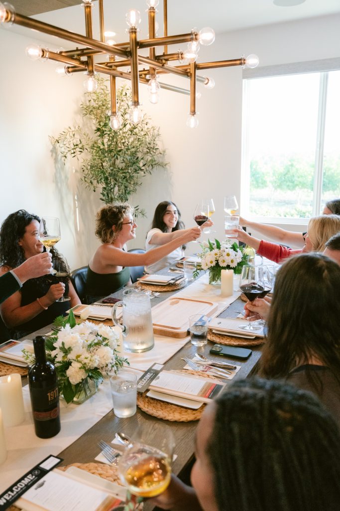 Editors raise their wine glasses in a toast around a beautifully set dining table during a CBC PR HOUSE Program dinner, surrounded by white floral centerpieces, branded place settings, and a wine bottle, beneath a gold industrial chandelier with greenery on the wall behind them.