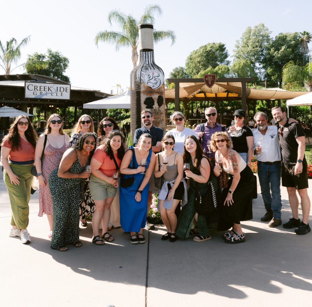 A group of approximately 18 editors and guests pose together in front of a large sculptural wine bottle at Wilson Creek Winery, palm trees and greenery behind them.