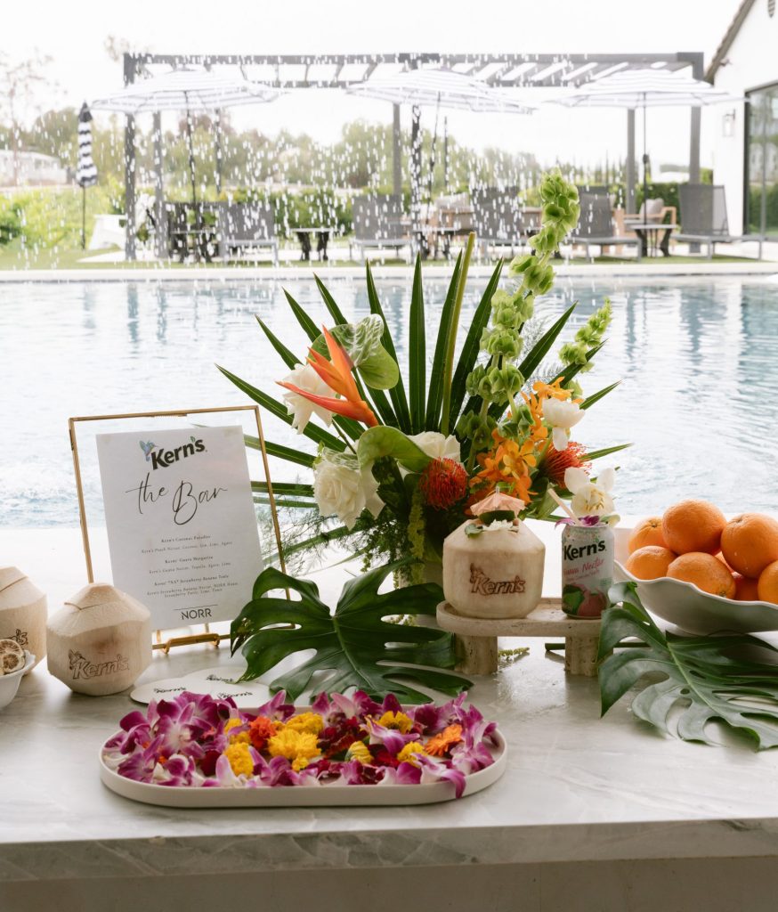A styled poolside bar setup for Kern's nectar drinks, featuring a "Kern's The Bar" menu sign, tropical floral arrangement, coconut cups, canned beverages, fresh oranges, edible flowers, and monstera leaves.