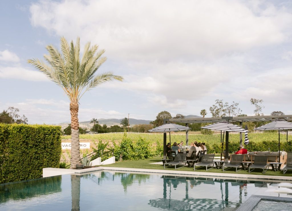 A serene infinity pool at a Temecula wine country estate reflects a lone palm tree and open sky, with editors gathered at a shaded outdoor table in the background.