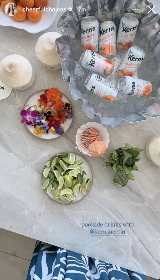 An Instagram Story from @cheerfulchoices captioned "poolside drinks with @kernsnectar" showing an iced bowl of Kern's Peach Nectar cans surrounded by edible flowers, lime wedges, and fresh mint on a marble surface.