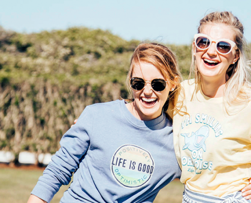Two women wearing Life Is Good t-shirt holding each other in sunlight